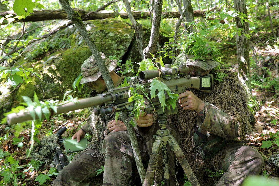 A soldier wearing  a camouflage uniform holds a weapon while another holds a notebook.