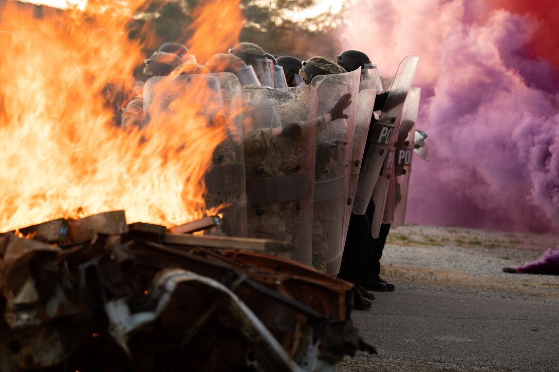 National Guardsmen and police in riot gear and holding shields stand in a line surrounded by flames and purple smoke.