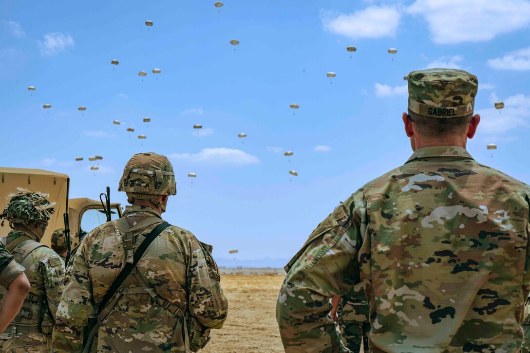 Soldiers watch as troops descend in the sky wearing parachutes.