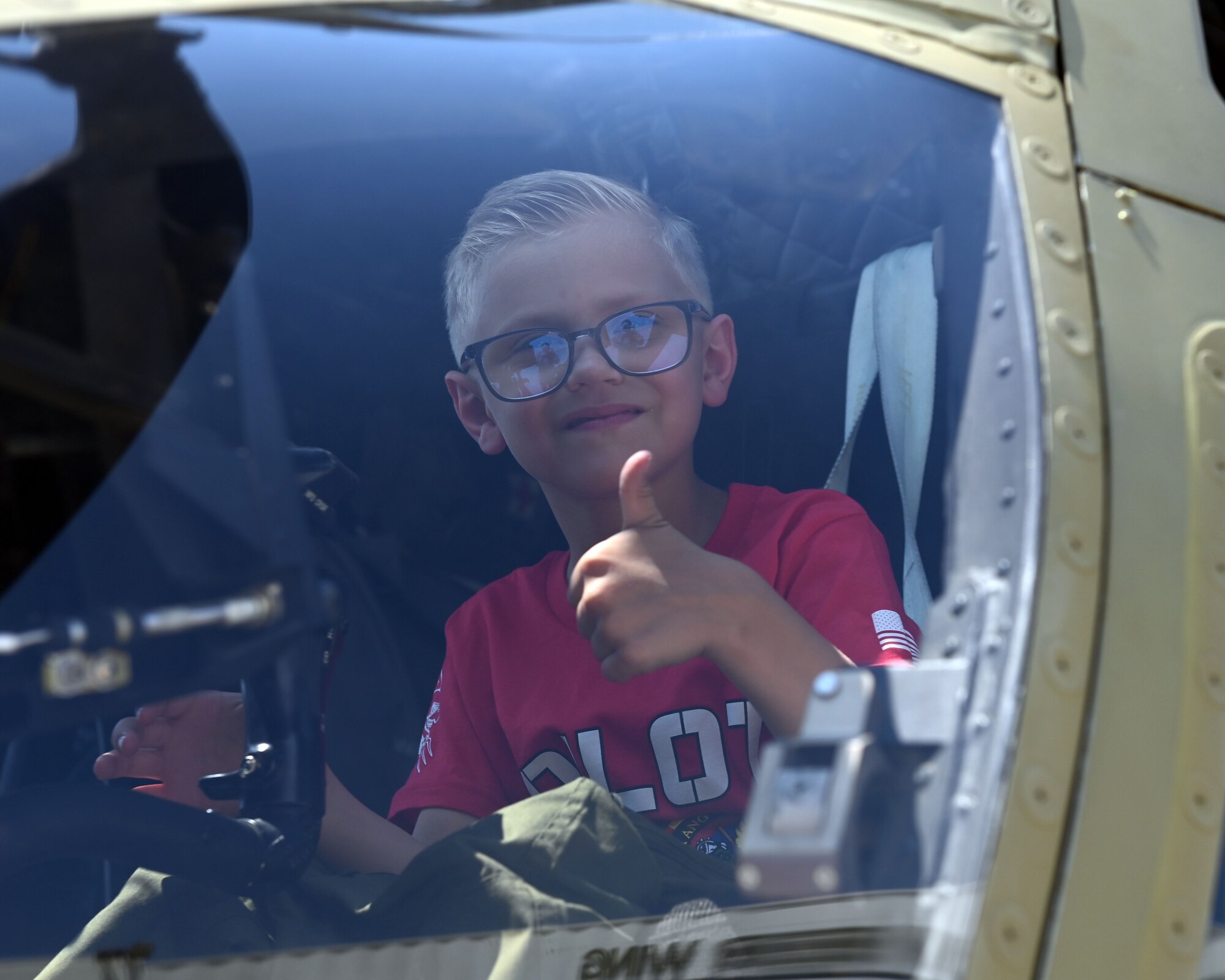Blonde boy with glass sits in the cockpit of a helicopter giving a thumbs up gesture.