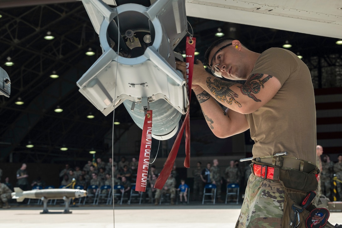 An airman wearing a toolbelt works on a weapon attached to an aircraft in front of an audience.