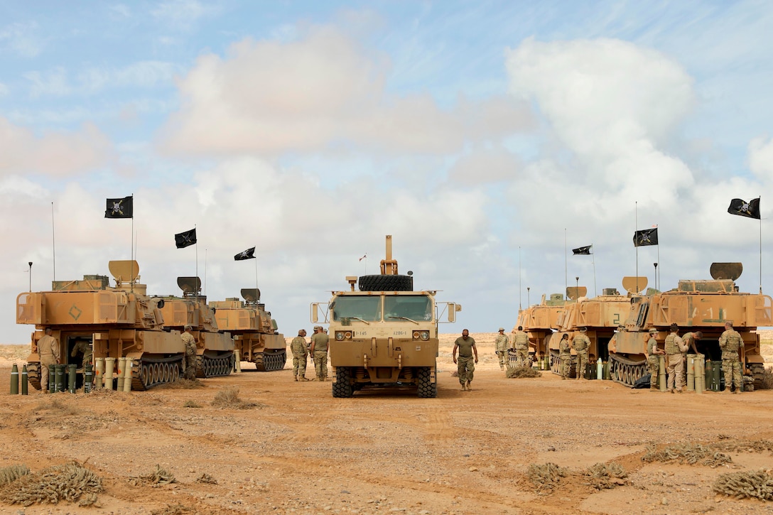 An armored vehicle sits in the middle of two rows of armored vehicles as guardsmen walk nearby in a desert-like area.