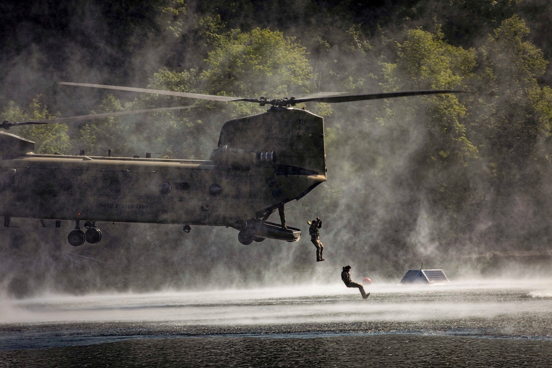 Two soldiers jump out of a helicopter hovering over a body of water with greenery in the background.