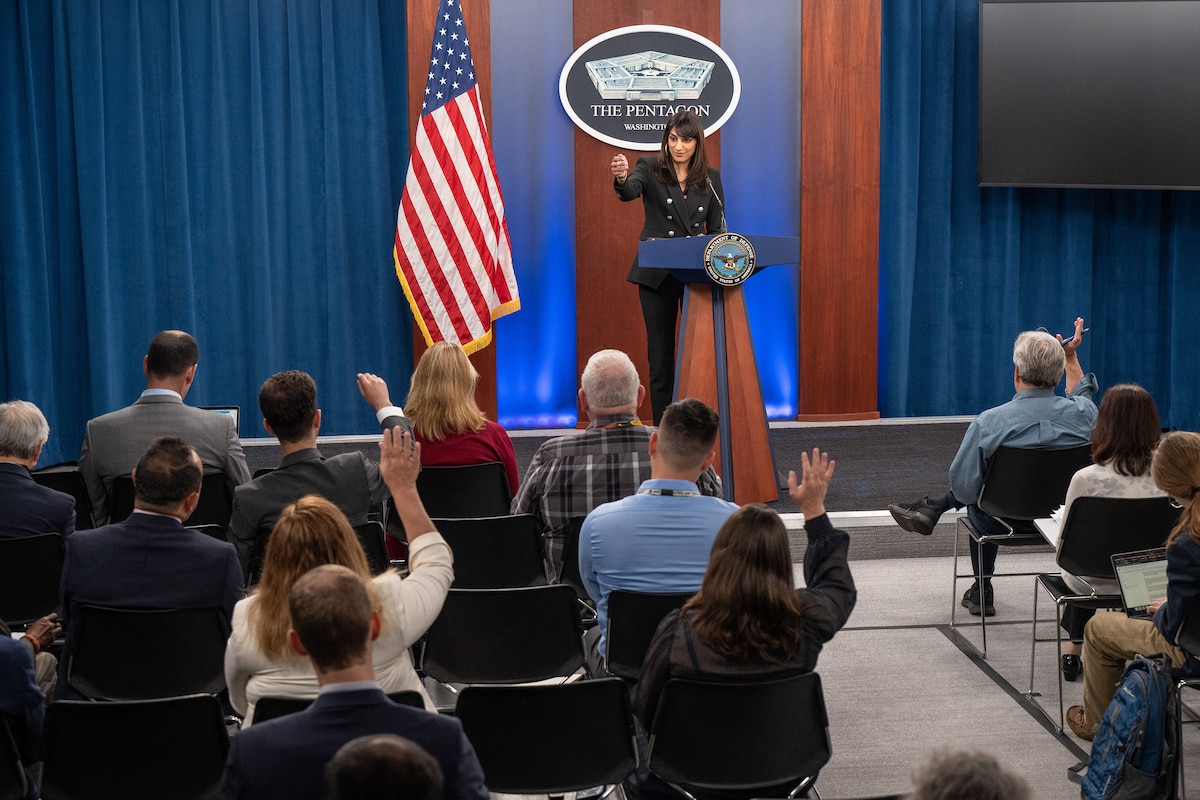 A civilian standing at a lectern gestures toward someone raising their hand in a seated audience.