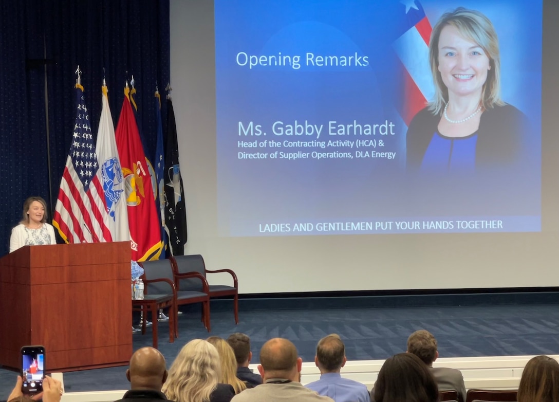 A woman speaking at a podium on a stage with a big screen behind her