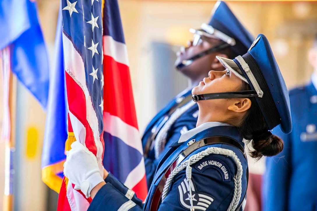 Two airmen stand side by side holding flags while looking up.