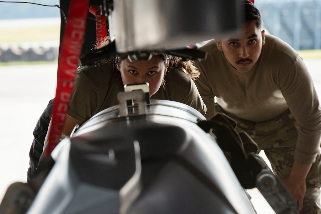 Two airmen standing next to each other crouch to look at the rearming area of an aircraft.