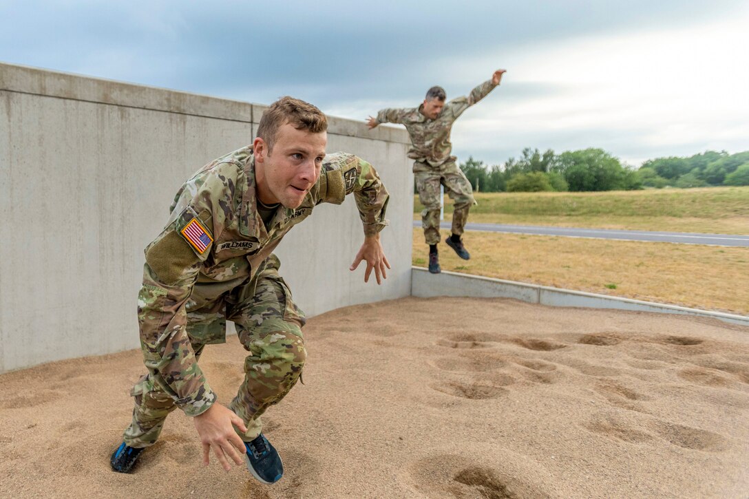 One soldier jumps from a concrete wall as another runs in the sand with greenery in the background.