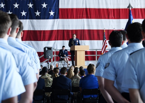 Uniformed man at podium in background speaks to people seated and in formation in the foreground