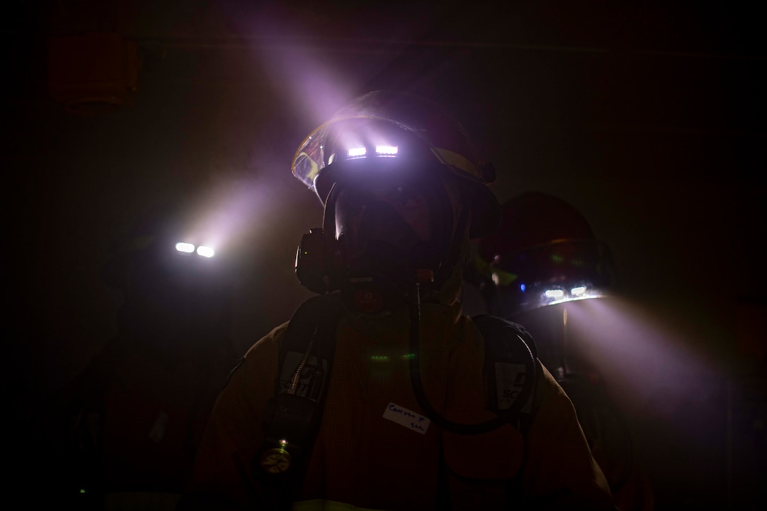 Sailors wearing firefighting gear are illuminated by helmet lights.