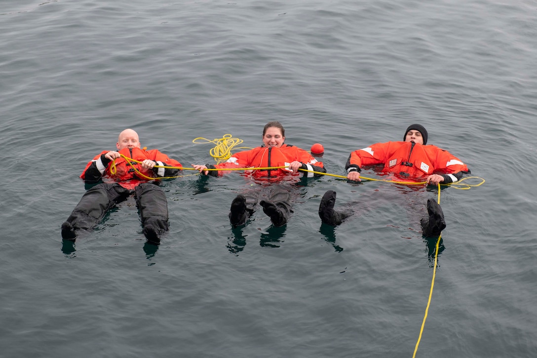 Three Coast Guardsmen in orange and black dry suits float on their backs while holding onto a yellow rope.