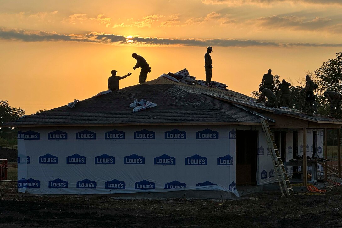 Airmen work on the roof of a house as the sun shines behind.