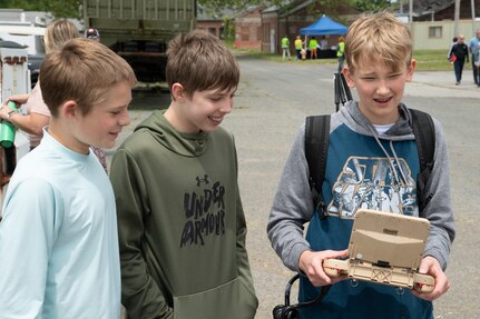 Three boys stand and one holds a controller for a semi-autonomous vehicle.