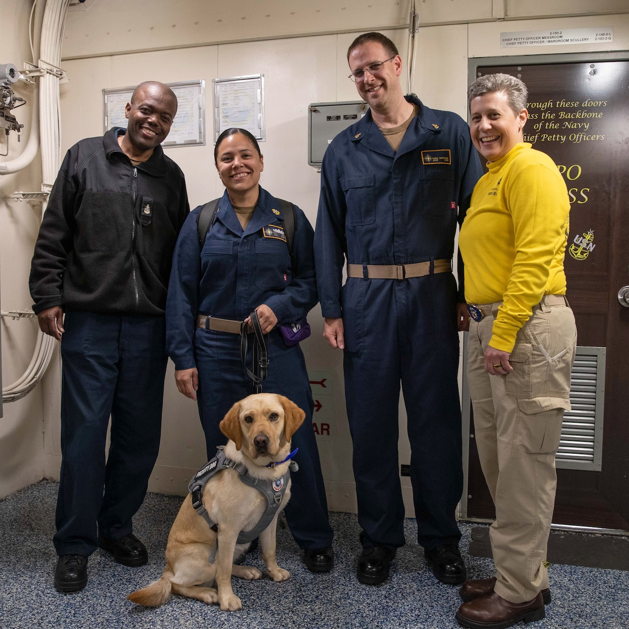 A dog sits in front of four smiling, standing sailors in a room.