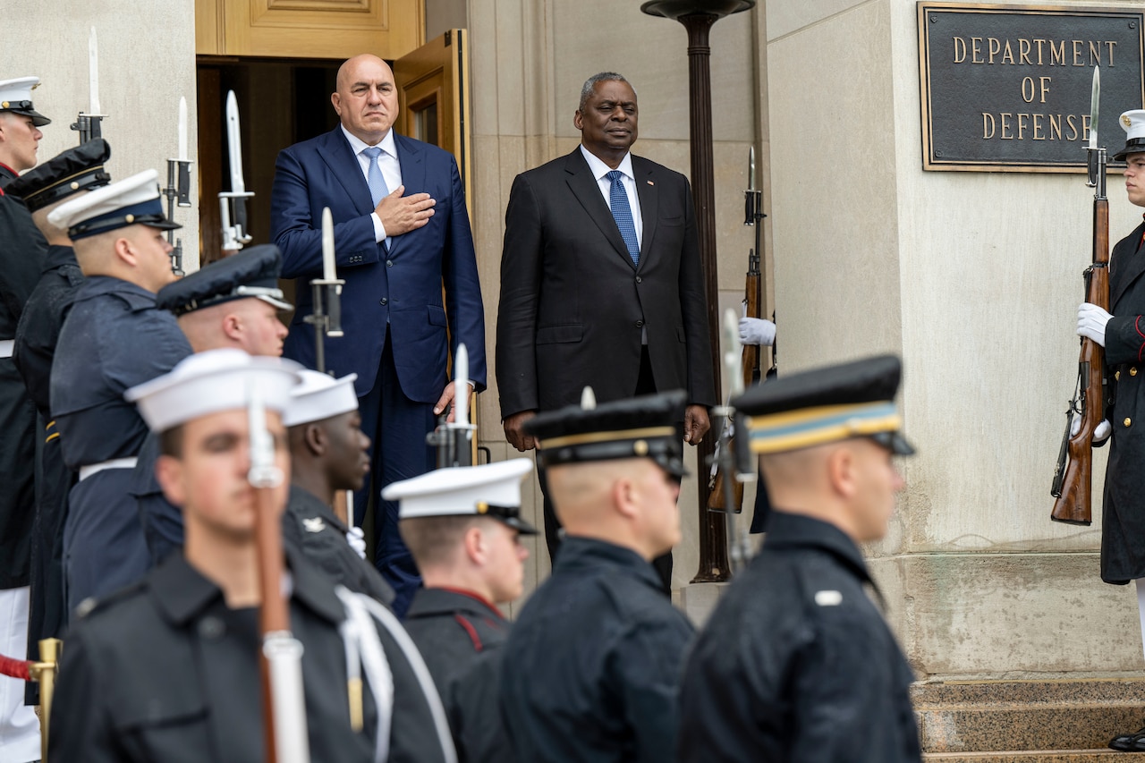 Two men dressed in business suits face forward at the top of an outdoor stairway. Service members line the stairs on each side.