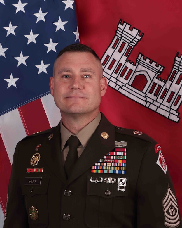 A man, Command Sergeant Major Douglas Galick, poses in front of a Red, White and Blue, United States Flag and a Red and Gold U.S. Army Corps of Engineers flag. He is wearing a in an\ black Army service dress uniform adorn with yellow stripes and medals.