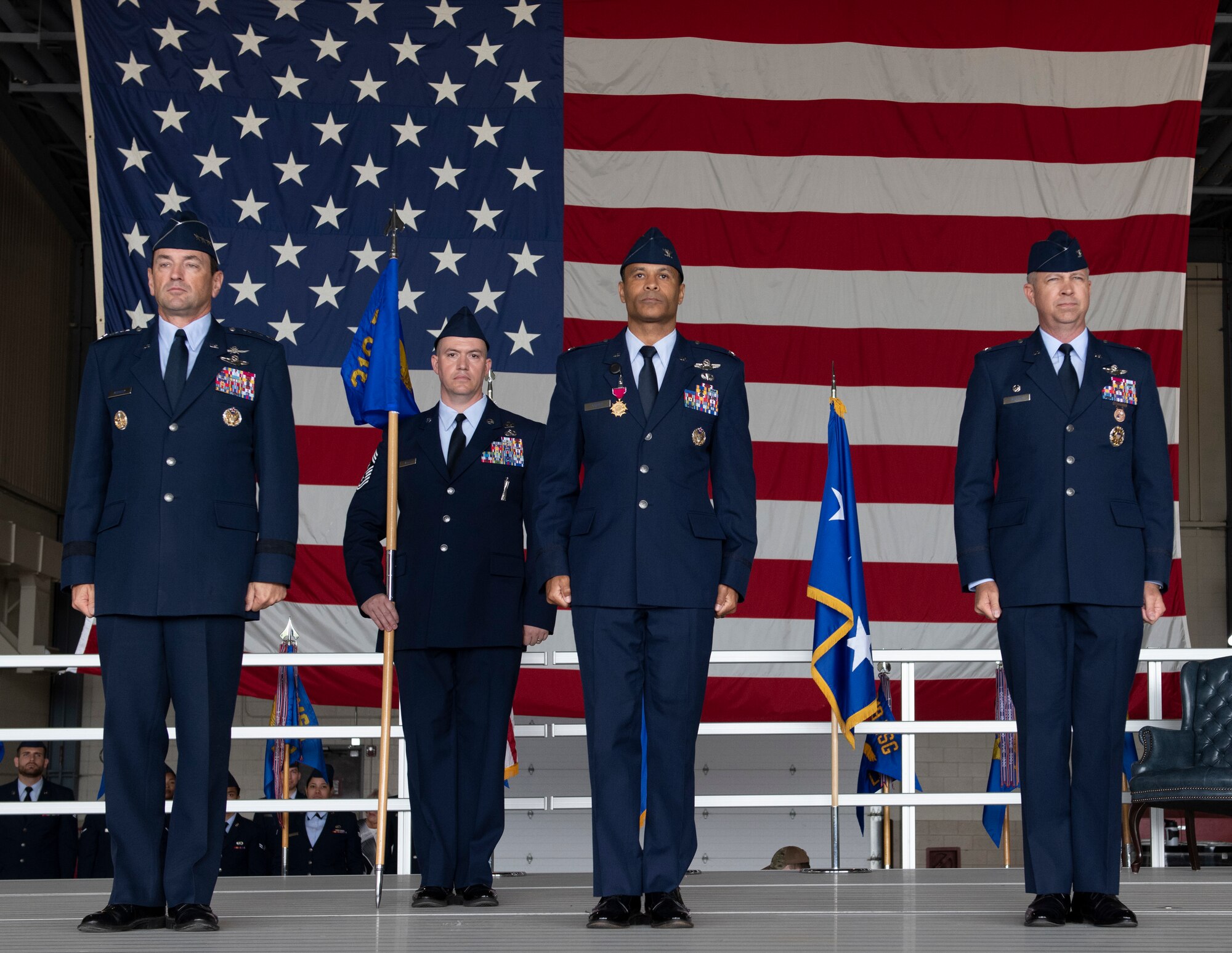 Four men in blue suits stand at attention in front of an American flag.