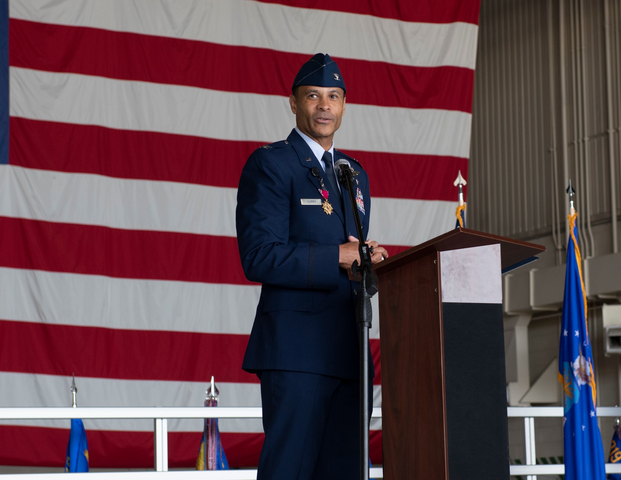 A man in a blue uniform speaks at a podium in front of an American flag.