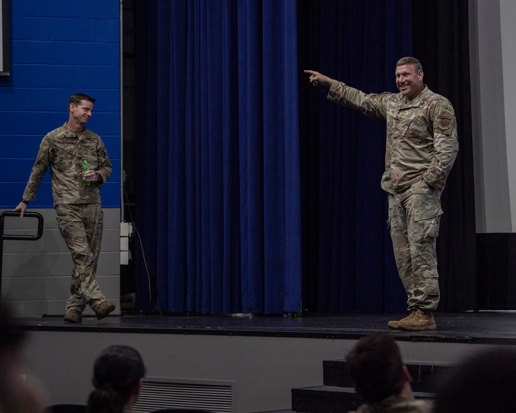 A photo of two men in uniform standing on a stage. The man on the right is pointing out into the crowd with a smile on his face.