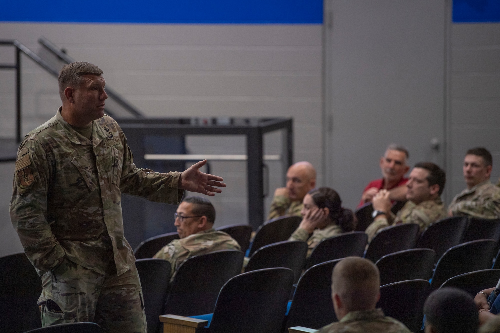 A photo of a man with his left hand extended, while talking to a crowd of people who are sitting in an auditorium.