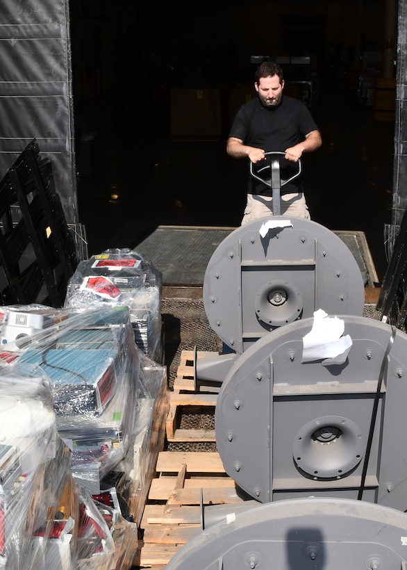 A man operates a pallet jack to unload large gray objects from the back  of a flat bad truck. The man and the items are outside but behind the man is a opening that leads into a dark warehouse.