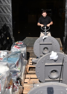 A man operates a pallet jack to unload large gray objects from the back  of a flat bad truck. The man and the items are outside but behind the man is a opening that leads into a dark warehouse.