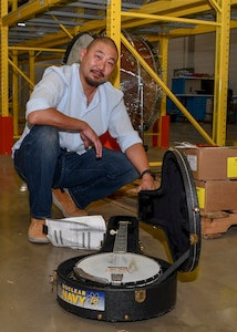 A man sits next to a banjo in a case on a warehouse floor.