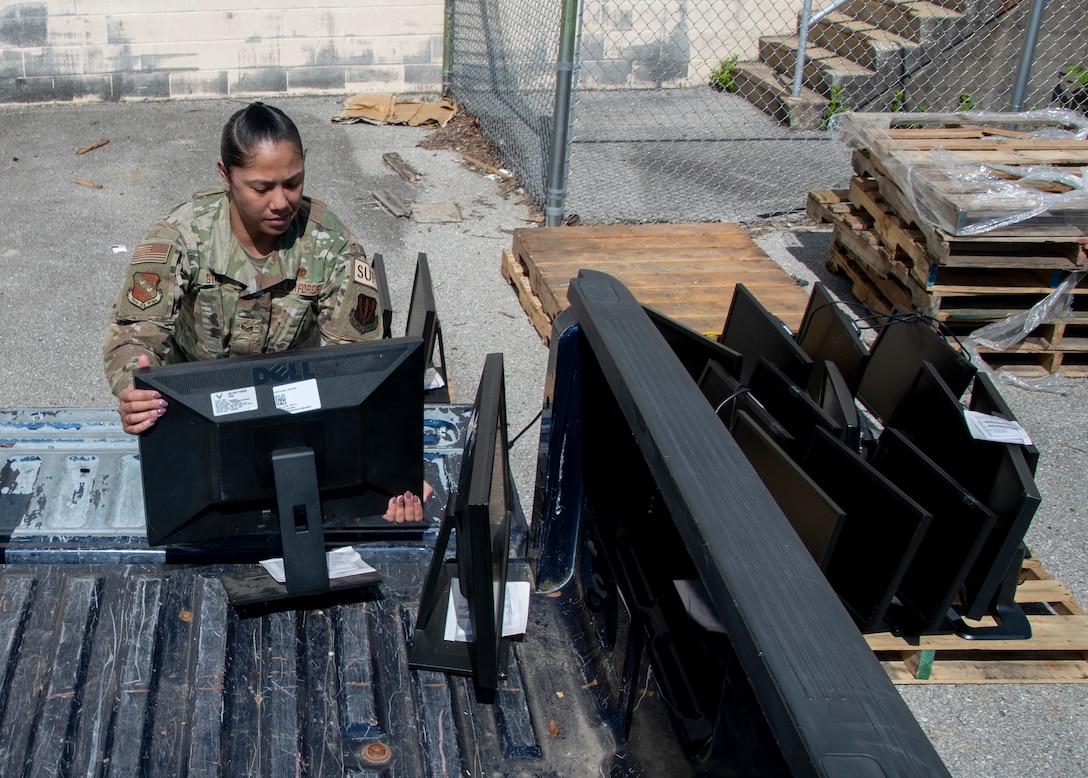 A woman in an Air Force uniform unloads computer monitors from the back of a pick up truck. She is outside in bright sun.