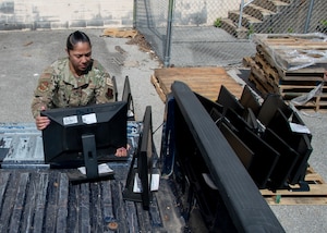 A woman in an Air Force uniform unloads computer monitors from the back of a pick up truck. She is outside in bright sun.