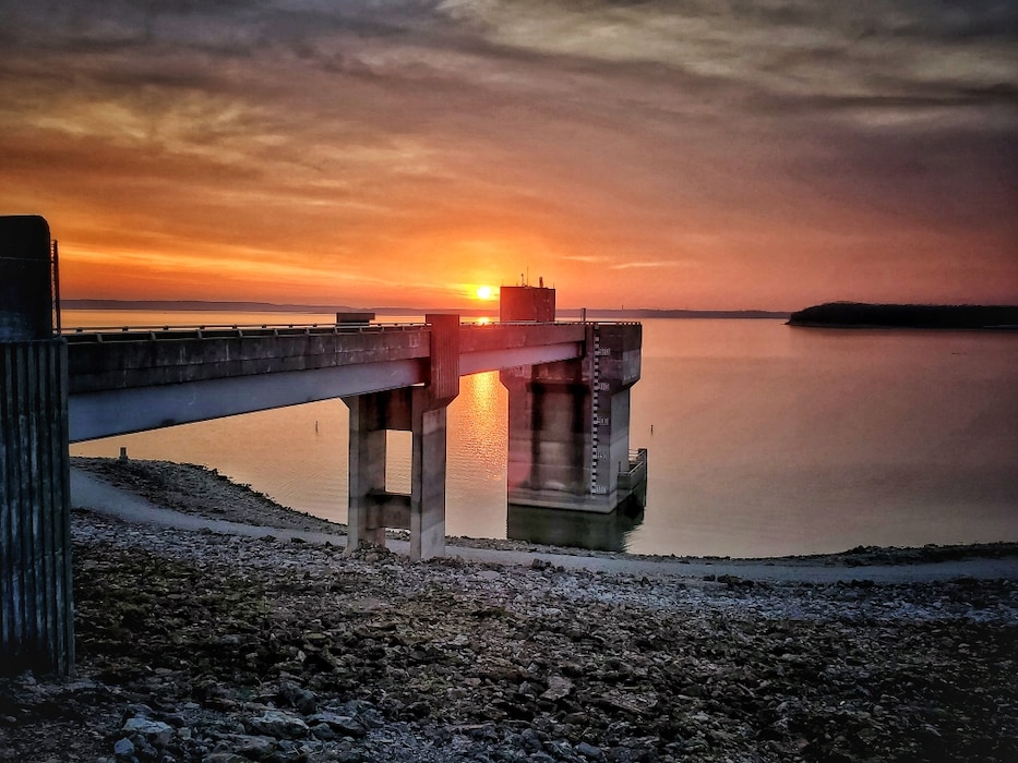 Clinton Lake can be seen with rock in the foreground, the dam in the middle of the photo, and a sunset in the background.