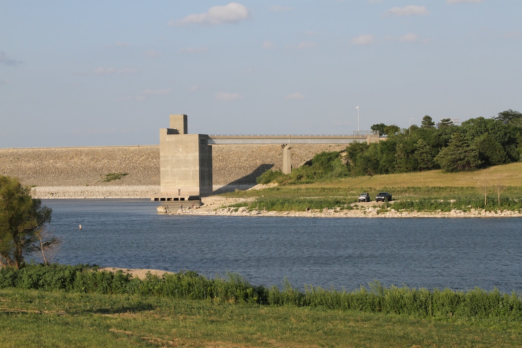 The dam at Milford Lake can be seen jutting into the water with grass in the foreground, trees to the left and right, and sky in the background.