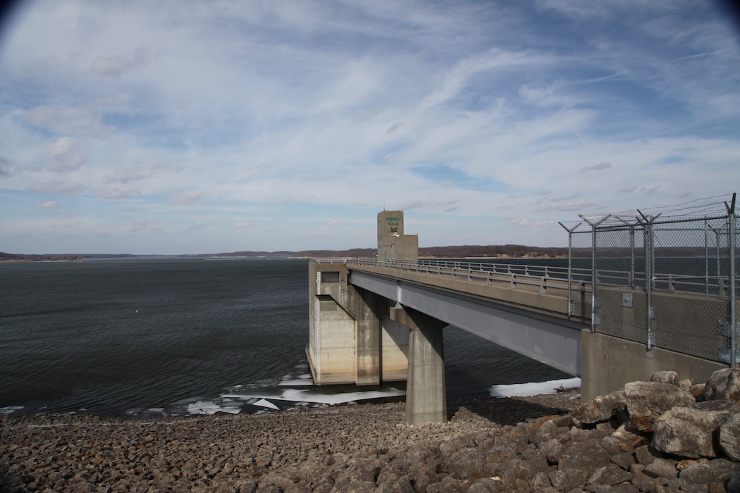 Perry Lake can be seen with the dam stretching into the water, rocks in the foreground and a cloudy sky in the background.