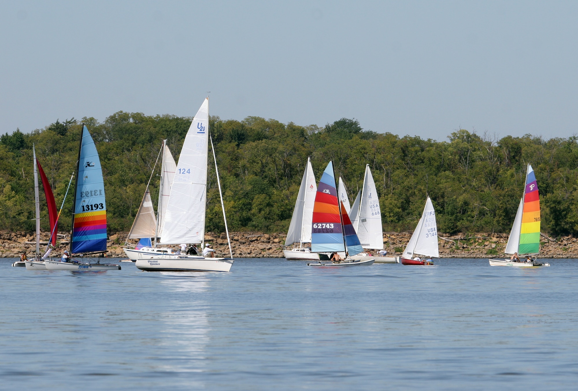 Several brightly colored sailboats can be seen on Perry Lake with water in the foreground and trees and sky in the background.