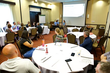 Photo taken from the back of a large hotel conference room. Military families face forward as presenters address the audience.