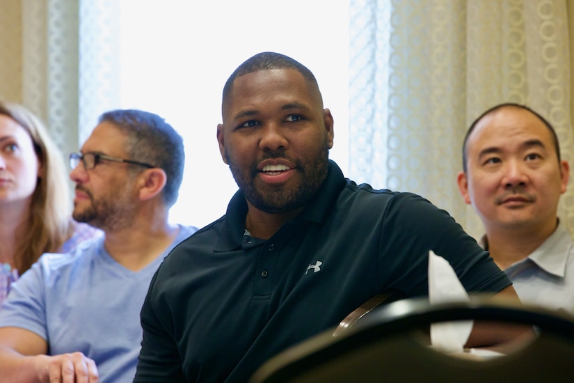 A man smiles while he listens to presenters speak.