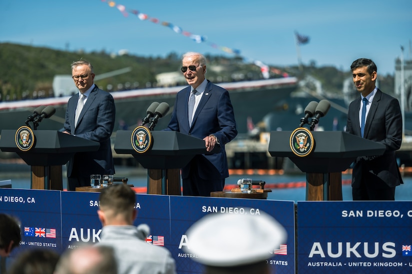 Three men in suits deliver remarks from an outdoor stage with a Navy ship in the background.