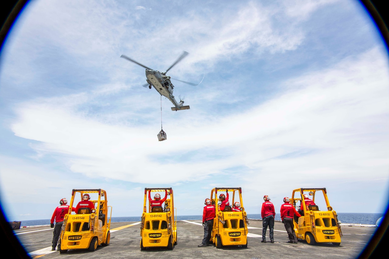 Sailors stand next to forklifts as a helicopter flies above with a cable dropping supplies.