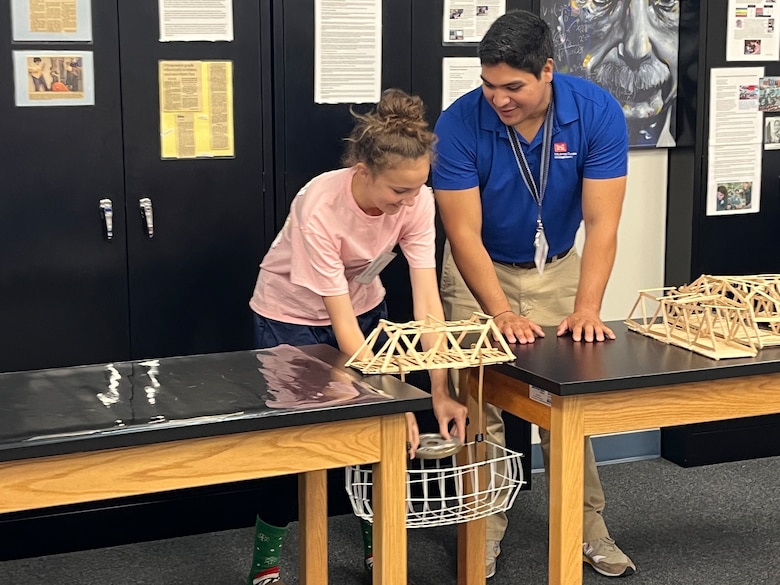Man and student putting weights in a basket to test popsicle bridge strength.
