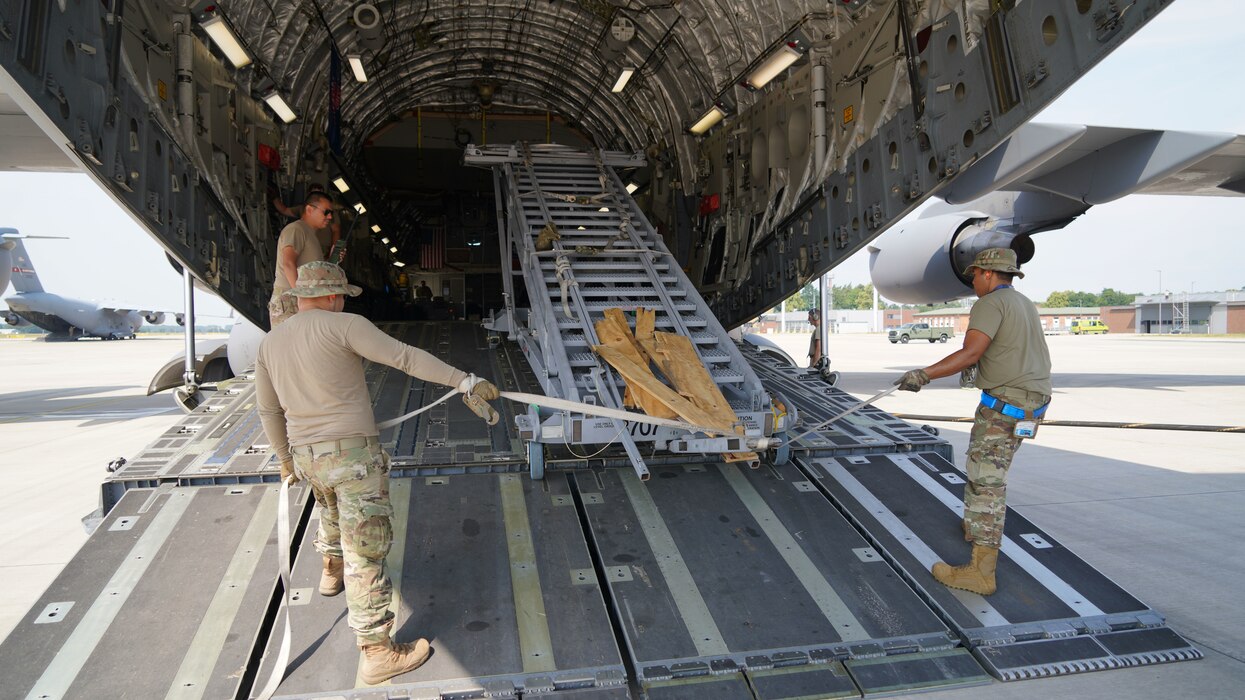 A photograph of three U.S. Air Force Airmen loading scaffolding equipment used by maintenance workers into the back of a military C-17. The Aircraft's ramp is down and the Airmen guiding the scaffolding are using straps to steer it into the aircraft.