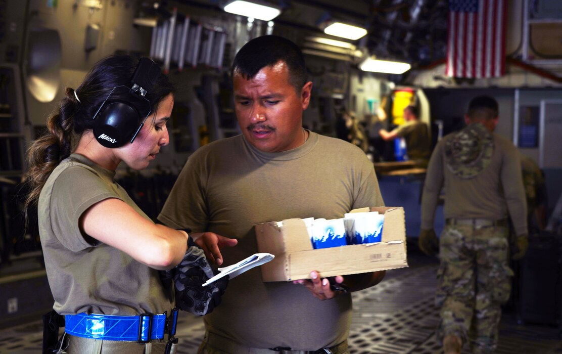 A female U.S. Air Force Airman reviews a document with another Male U.S. Air Force Airman inside the cargo hold of a military C-17. The inside of the C-17 is dimly lit with a small amount of natural sunlight is falling onto both the Airmen as it enters from behind the aircraft. Two additional Airmen can be seen working inside the aircraft behind the previous Airmen mentioned with a U.S. flag hung above them.