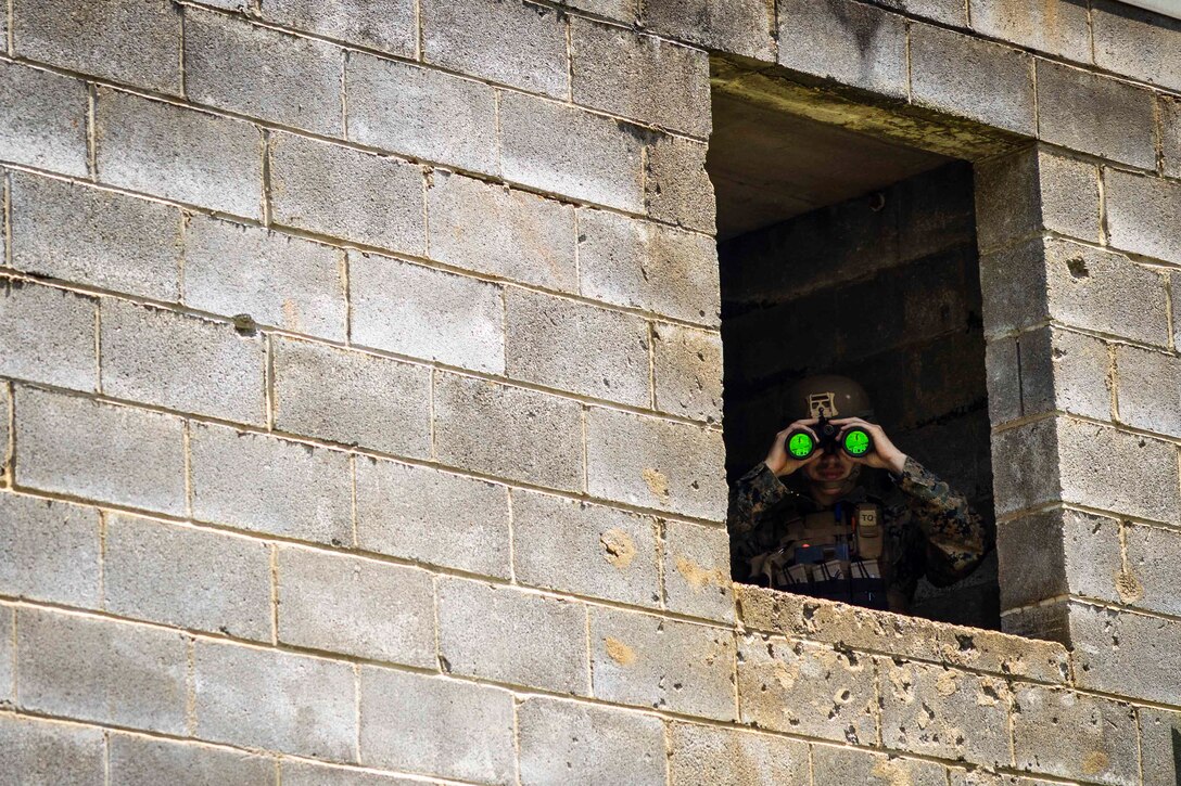 A Marine uses binoculars to look out a window.