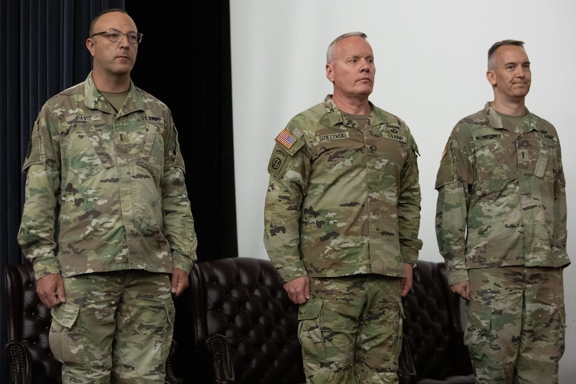 Three Army officers stand at attention on a stage.