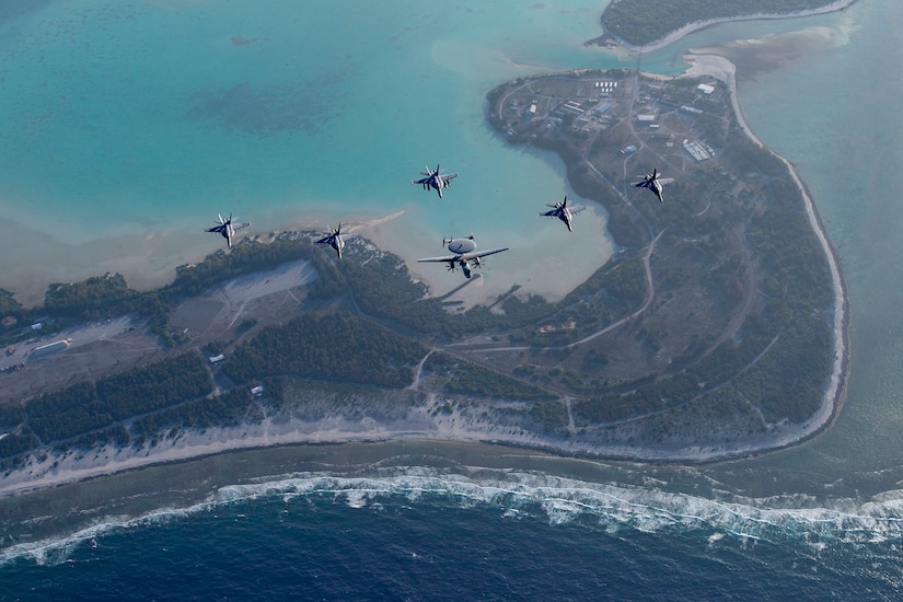 Six Navy aircraft fly over the coastline of an island in the Pacific Ocean.