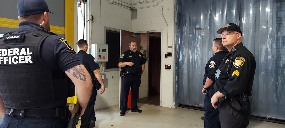 Several men dressed in dark colored police uniforms stand around and chat in a loading dock area of a building.