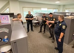 Five men in dark colored police uniforms with various protective equipment stand in the middle of a few office cubicles. A window looking outside is in the background. The room has white walls.