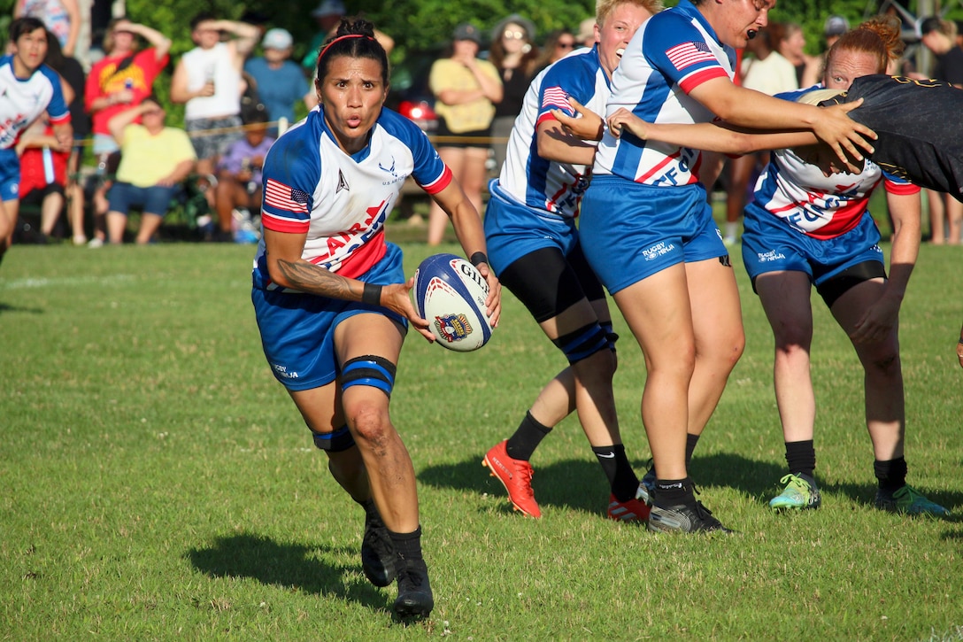 An airman runs with a ball during a rugby match as team members block an opposing player in the background.