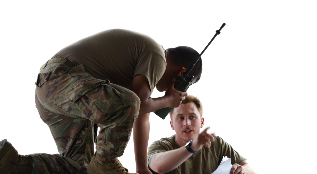 A U.S. Air Force Airman kneeling down on the edge of the ramp door inside a military C-17 aircraft listening to a radio while another U.S. Air Force Airman is pointing towards the inside of the cargo area.