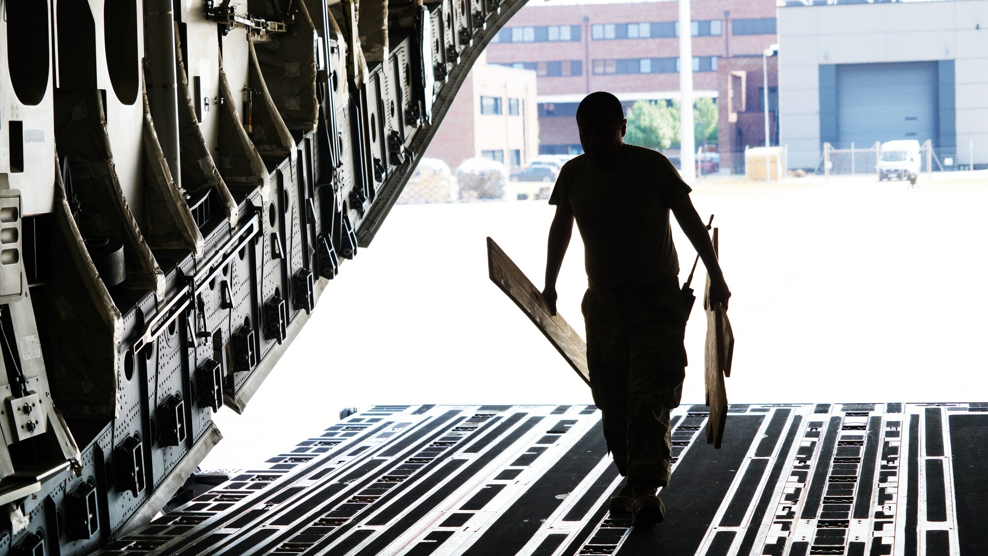 A photograph silhouette looking towards and out the back of a military C-17 aircraft as a U.S. Air Force Airman carrying two pieces of equipment walks up the ramp.