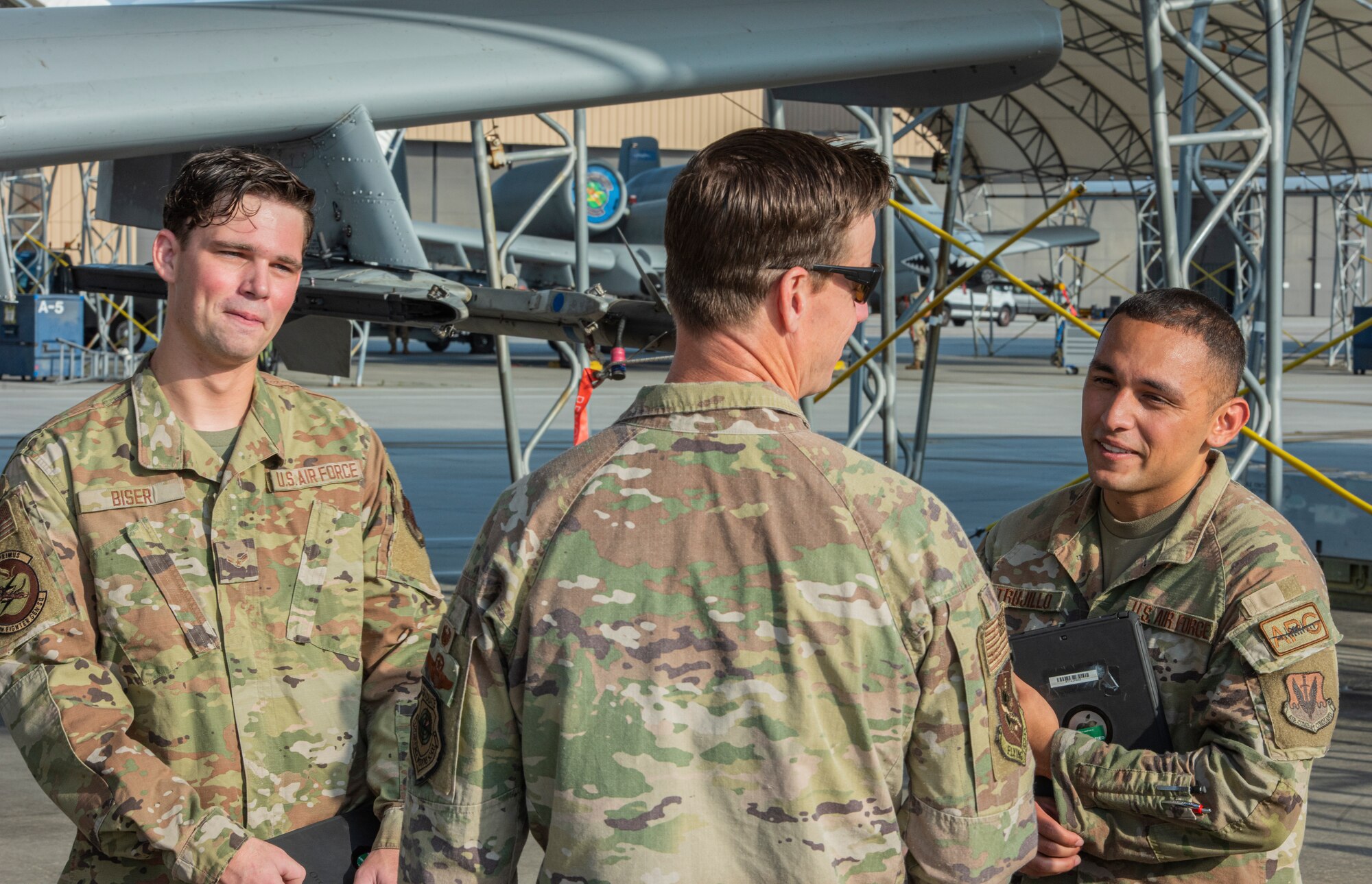 A photo of three people standing on a flight line with A-10C Thunderbolt II aircraft in the back ground, talking.