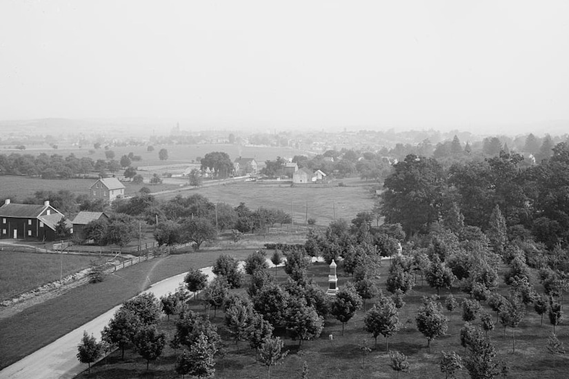 A view of a rural landscape, complete with trees, a few houses and a few monuments.